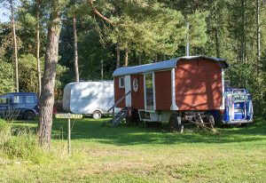 Roter Bauwagen mit großem Fenster auf einer Wiese. Daneben einige Autos und Caravans.